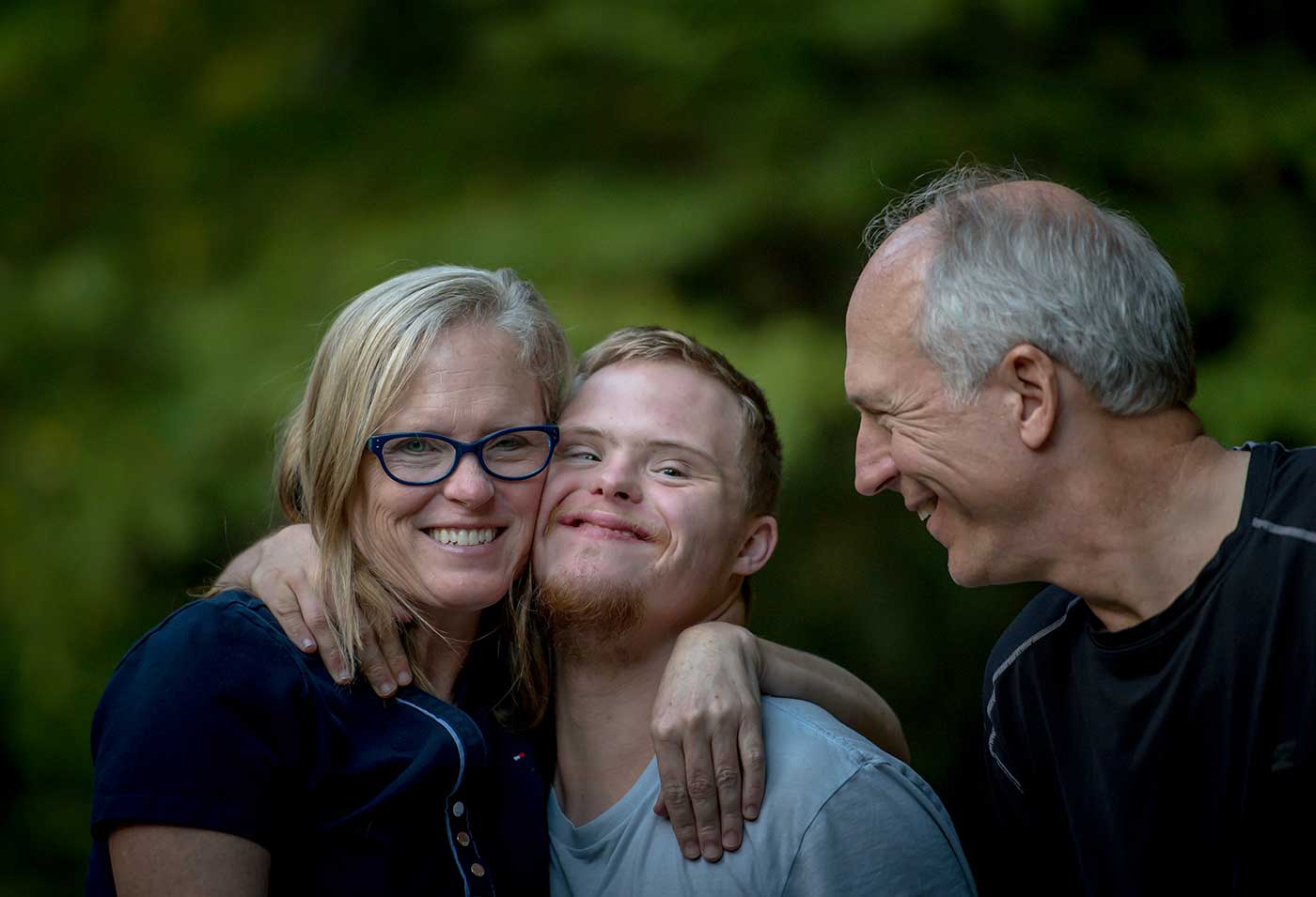 Smiling mom and dad hugging their son with Down's Syndrome, with the mom and son looking at the camera and the father looking at the son.